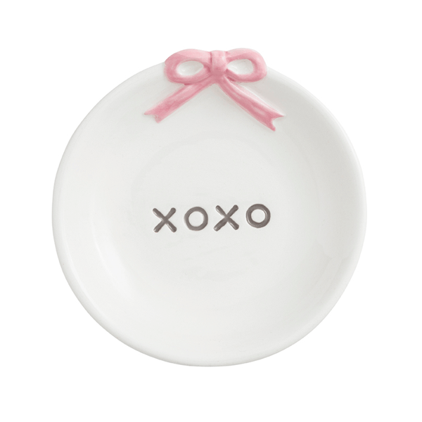 A white ceramic ring holder shaped like a shallow dish, featuring a pink ribbon bow at the top and the word 'XOXO' printed in gray in the center.