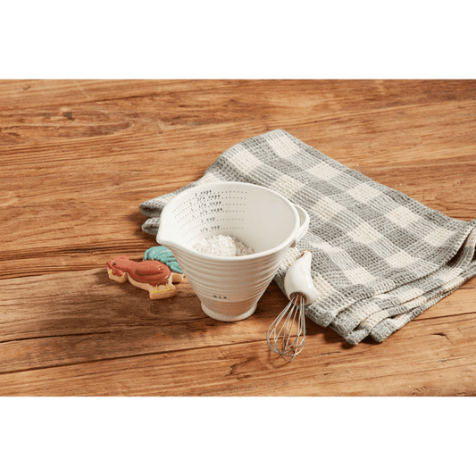 A white ceramic measuring cup filled with flour, placed on a rustic wooden table alongside a gray-and-white checkered cloth, a metal whisk, and small animal-shaped cookies.
