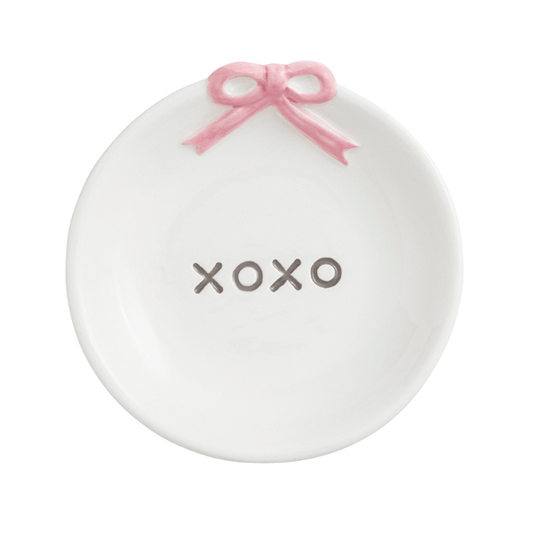 A white ceramic ring holder shaped like a shallow dish, featuring a pink ribbon bow at the top and the word 'XOXO' printed in gray in the center.
